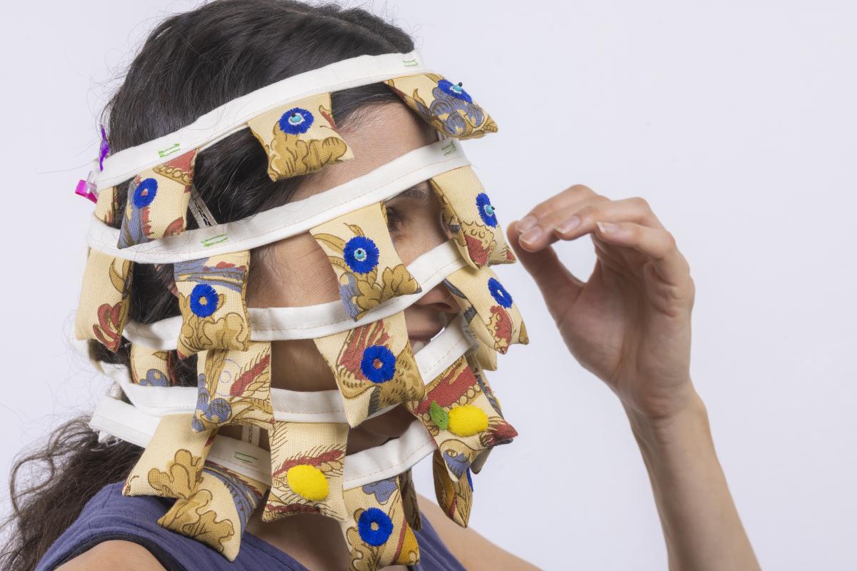 A headshot of a person viewed from the side, wearing pieces of cloth with vertical fabric pieces in horizontal rows overlapping their face, as they look to the right.