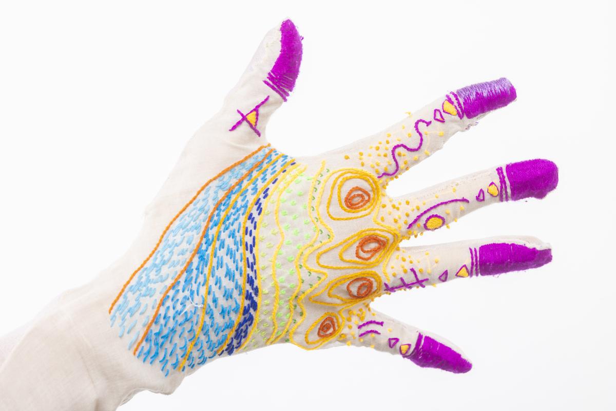 A hand showing towards the camera with all 5 fingers stretched. The hand has multicolored cloth and fabric outlining certain aspects of the fingers and hand in a beautiful array of colors.