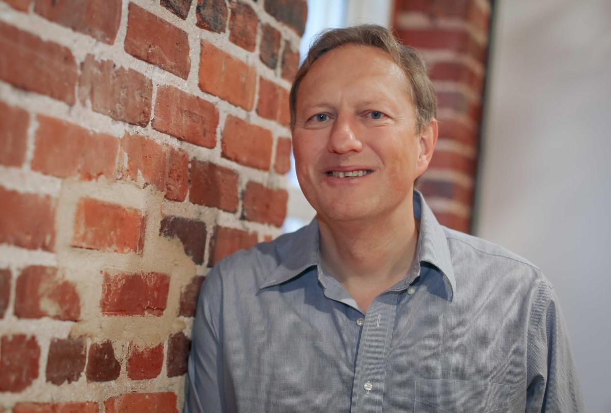 Johan Rempel, wearing a blue collared shirt, stands facing the camera next to a brick wall indoors.